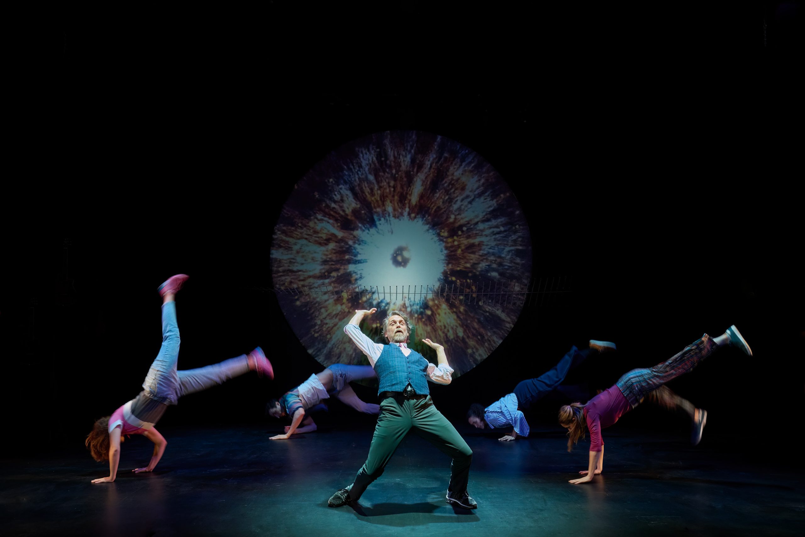 Four performers from Theatre Gargantua balance on their hands in synchronized donkey-kick poses, with Michael Gordon Spence leaning back as a burst of projected light forms a sound-wave pattern behind them.