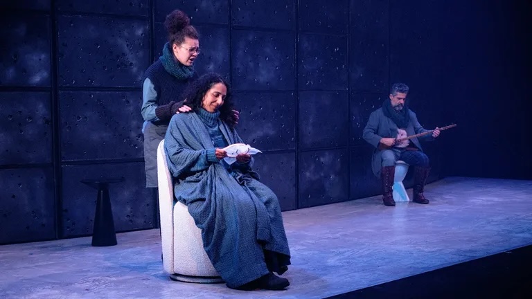 A weary mother sits slumped in a wooden chair while Martha leans over her with fierce intensity, and in the background the silent old man sits on a stool with his mandolin, watching.