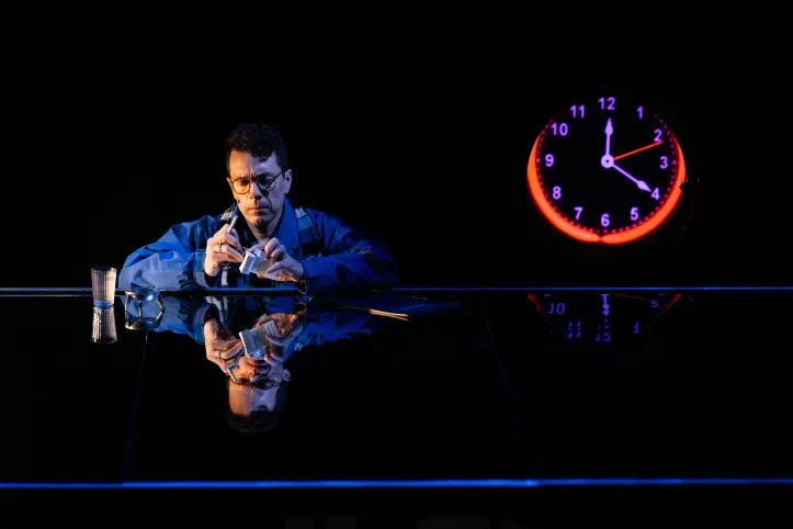 Olivier Normand sits at a small table in a dim, orange glow, his mirrored reflection beside him. He leans forward with a cigarette in one hand and a drink in the other, framed by the familiar porthole that now appears as a luminous orange clock. The surface of the reflection blurs the boundary between the two brothers, capturing the production’s mix of domestic detail and cosmic longing.
