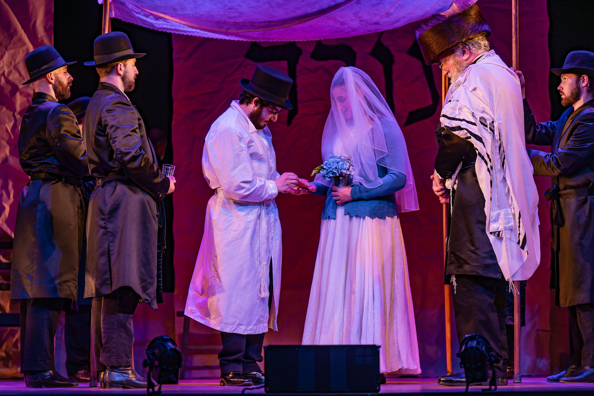 Jewish wedding under the canopy, bride and groom both dressed in white, rabbi andd two witnesses standing to th siide.